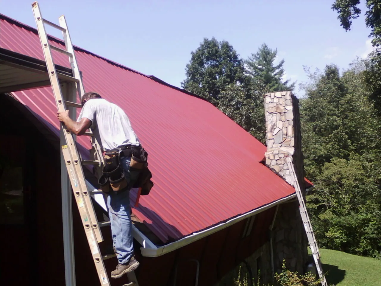Roofer on a ladder beside a red metal roof and stone chimney — Soffit & Fascia Repair by Roofing Brothers in Rossiter